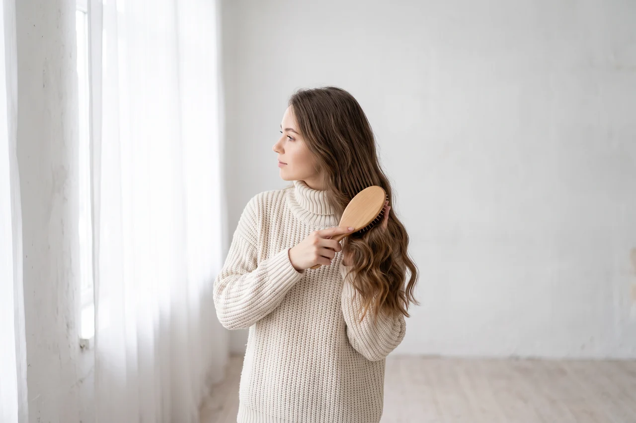 Una ragazza con un maglione color crema si pettina i capelli lunghi con una spazzola di legno, vicino a una finestra luminosa