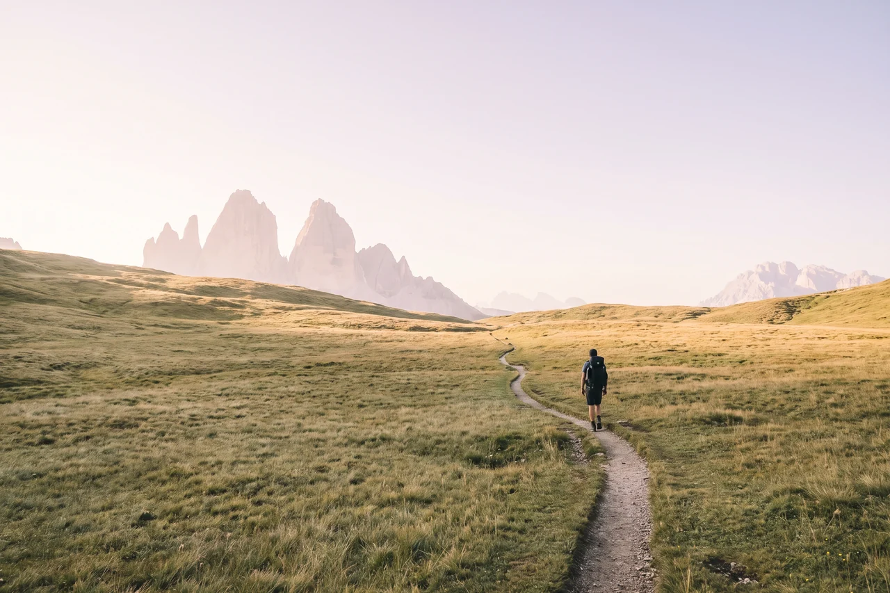 Un escursionista con lo zaino cammina su un sentiero tra i prati verso le cime delle Dolomiti