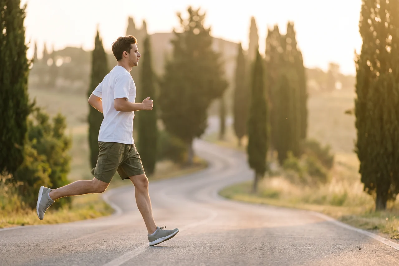 Un ragazzo con maglietta e pantaloncini corre da solo su una strada di campagna all’alba, tra le colline e i cipressi